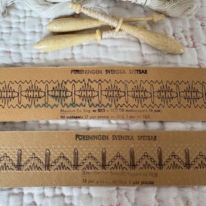 May include: A top-down view shows antique lace-making tools. Two wooden bobbins with white thread rest above two brown paper strips with detailed black lace designs and text. White thread spools are visible.