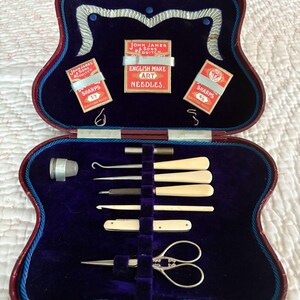 May include: An open, antique sewing kit with a red and blue case. Inside, a deep blue velvet lining holds various sewing tools, including needles, scissors, and bone-handled implements. The kit also contains small boxes of "JOHN JAMES & SONS REDDITCH" needles.