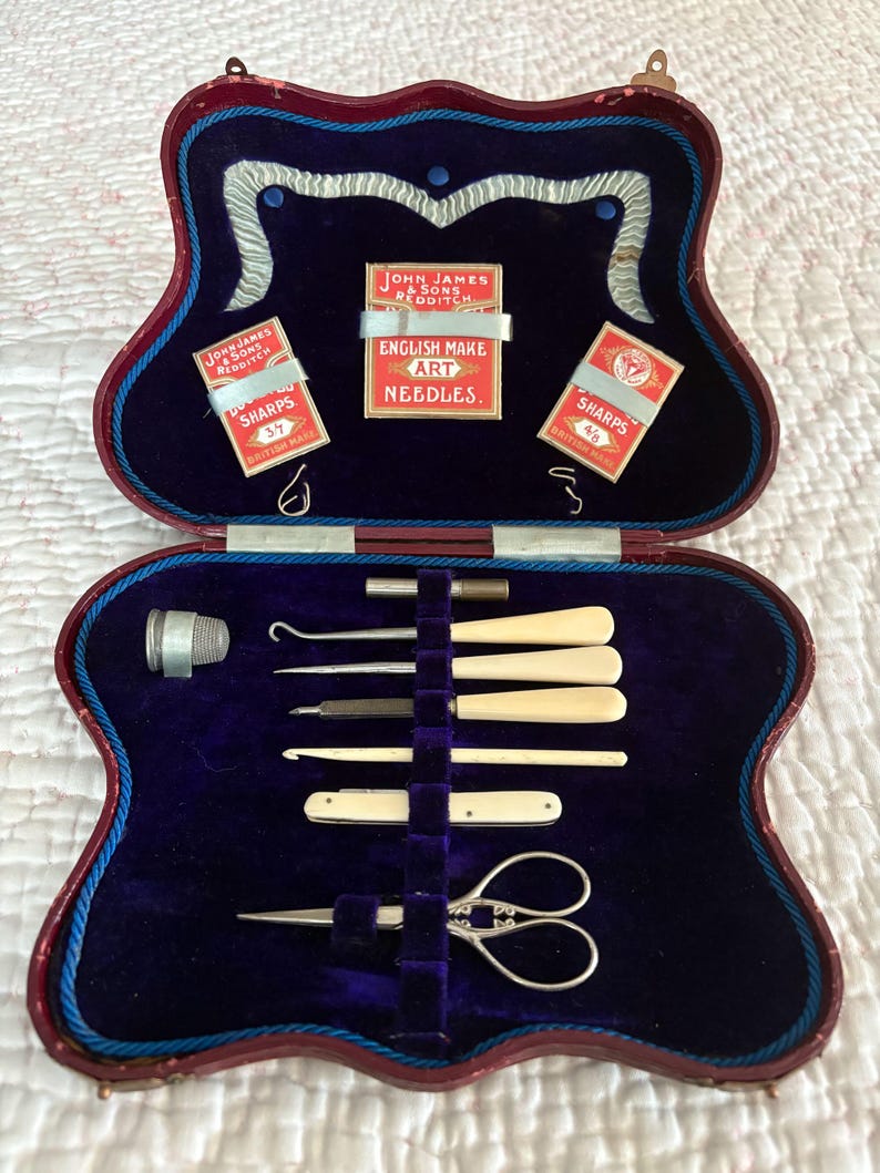 May include: An open, antique sewing kit with a red and blue case. Inside, a deep blue velvet lining holds various sewing tools, including needles, scissors, and bone-handled implements. The kit also contains small boxes of "JOHN JAMES & SONS REDDITCH" needles.
