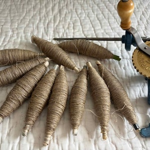 May include: A collection of eleven light brown, spindle-shaped bobbins wrapped in natural fibre thread, positioned next to a vintage metal yarn winder featuring a gold-coloured gear with visible text and a blue clamp mechanism.