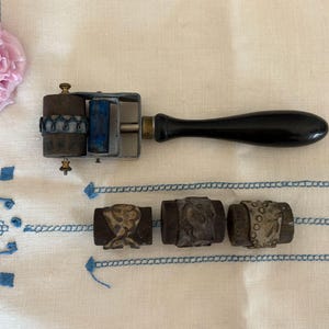 May include: A vintage printing set comprising a roller with a black handle and three interchangeable stamps. The set is made of metal and wood, with a retro aesthetic. A pink flower and embroidered fabric complete the scene.
