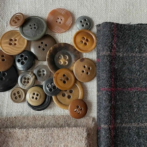 May include: A collection of various-sized buttons in shades of brown, black, and gray, with four-hole and two-hole designs. A piece of gray plaid fabric with red accents and a piece of tan fabric are also present, suggesting a sewing or crafting theme.
