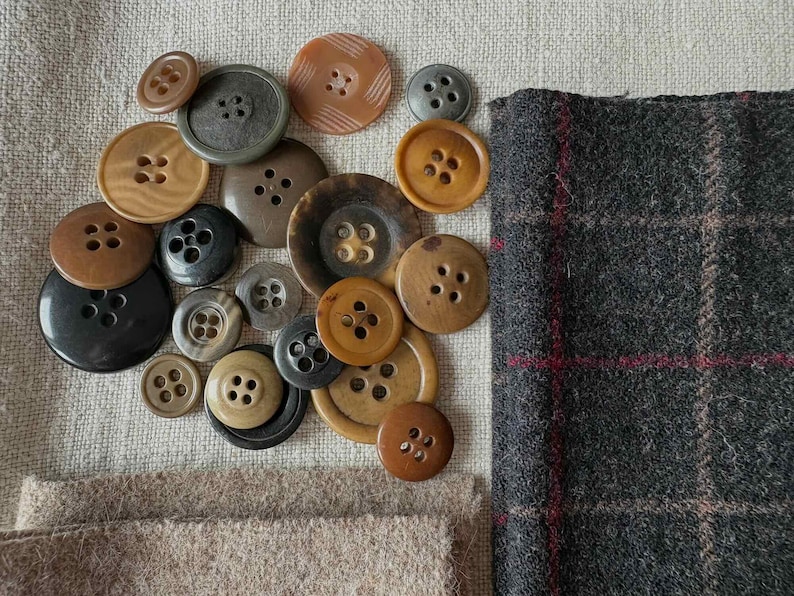 May include: A collection of various-sized buttons in shades of brown, black, and gray, with four-hole and two-hole designs. A piece of gray plaid fabric with red accents and a piece of tan fabric are also present, suggesting a sewing or crafting theme.