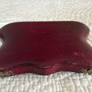 May include: A burgundy-colored, antique case with a curved, irregular shape. The case has a textured surface and is secured with small, ornate metal clasps. The edges show signs of wear, suggesting its age and use.