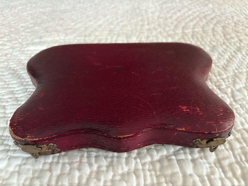 May include: A burgundy-colored, antique case with a curved, irregular shape. The case has a textured surface and is secured with small, ornate metal clasps. The edges show signs of wear, suggesting its age and use.