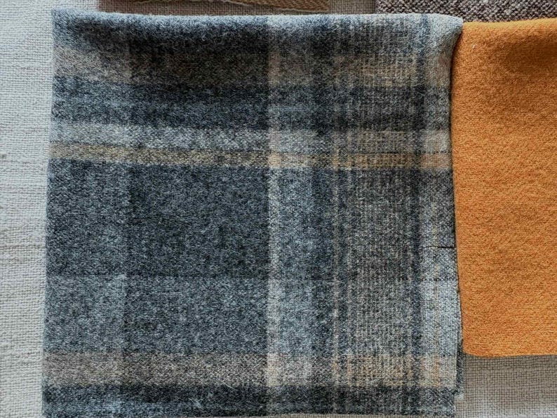 May include: Close-up of fabric swatches. The main swatch is a blue and gray plaid pattern. Adjacent is a solid orange swatch. The fabrics appear to be wool or a similar textured material, suitable for apparel or home decor.