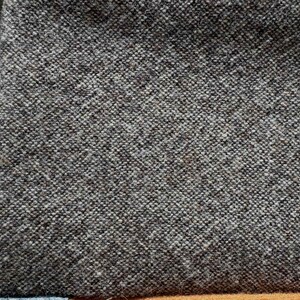 May include: Close-up of a textured, dark gray fabric with a woven pattern. The fabric has a slightly rough appearance and is likely a type of tweed or wool. Other fabric samples are visible.