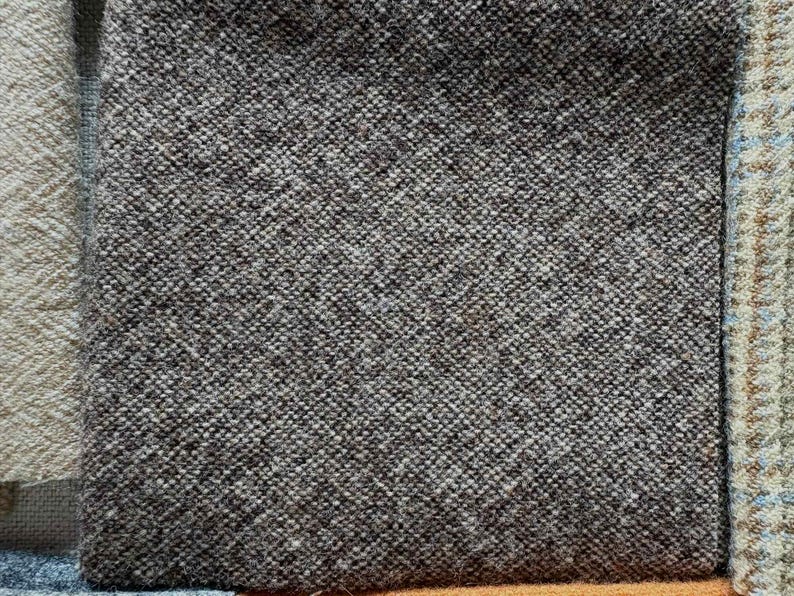 May include: Close-up of a textured, dark gray fabric with a woven pattern. The fabric has a slightly rough appearance and is likely a type of tweed or wool. Other fabric samples are visible.