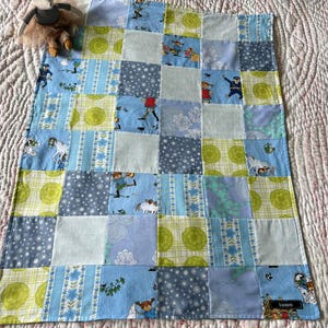 May include: A rectangular patchwork quilt featuring a grid of fabric squares in shades of light blue, lime green, and white. The fabric patterns include cartoon characters, floral motifs, polka dots, and plaid designs, finished with a small black label.