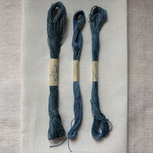 May include: Three skeins of blue embroidery floss are shown against a white fabric background. Each skein is wrapped with a paper label featuring a decorative design. The floss has a slightly textured appearance.