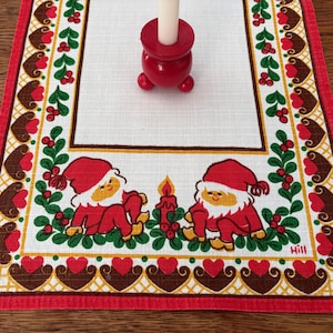 Peut inclure: Un chemin de table rectangulaire blanc avec une bordure rouge et un motif répétitif de cœurs marron et de treillis jaune. Le centre présente deux personnages illustrés en chapeaux et vêtements rouges assis près d'une bougie, avec le nom Hill.