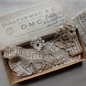 May include: An open cardboard box containing antique crochet lace and trim. The box is labeled "DOLLFUS-MIEG & C" and "DMC". The lace is beige and features various patterns and floral accents. A piece of lace is outside the box.