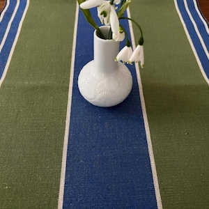 May include: A striped table runner in shades of blue, green, and white. A small white vase with a floral pattern contains white flowers. The runner is made of a textured fabric, suitable for a dining table.