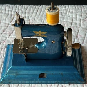 May include: A vintage blue sewing machine with a gold eagle emblem. The machine has a silver needle and a wooden handle. A spool of yellow thread sits on top. The machine is on a blue base.
