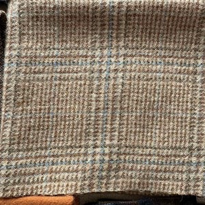May include: Close-up of a tan and brown plaid fabric with blue accents. The fabric has a textured appearance and a classic pattern. The image shows a portion of the fabric, highlighting its weave and color variations. The fabric is likely used for clothing or upholstery.