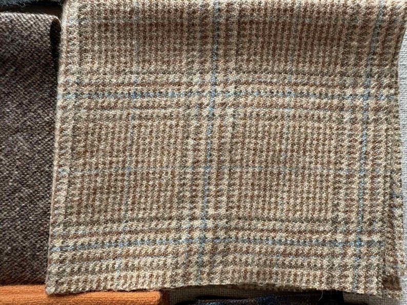 May include: Close-up of a tan and brown plaid fabric with blue accents. The fabric has a textured appearance and a classic pattern. The image shows a portion of the fabric, highlighting its weave and color variations. The fabric is likely used for clothing or upholstery.