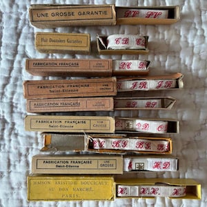 May include: A collection of vintage French ribbon spools in open cardboard boxes. The boxes are labeled with French text, including "Fabrication Française" and "Une Grosse Garantie." The ribbons are white with red lettering.