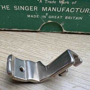May include: A silver-colored sewing machine presser foot. The presser foot is made of metal and has a curved shape with a hole and a small hook. The text "THE SINGER MANUFACTURING MADE IN GREAT BRITAIN" is visible in the background.