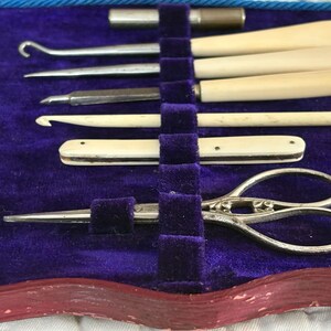 May include: A vintage sewing kit with various tools, including ivory-handled hooks, a small pair of scissors, and other implements. The tools are arranged in a purple velvet-lined case with a red border.