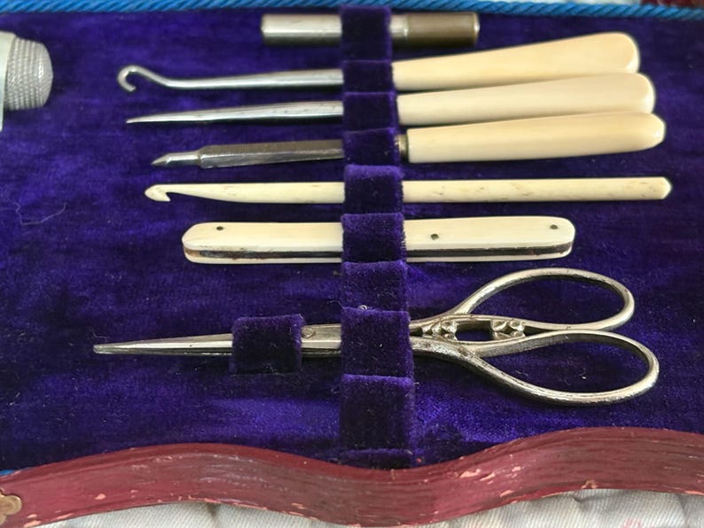 May include: A vintage sewing kit with various tools, including ivory-handled hooks, a small pair of scissors, and other implements. The tools are arranged in a purple velvet-lined case with a red border.