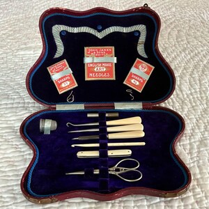 May include: An open antique sewing kit with a burgundy, scalloped case and a deep blue velvet interior. The kit contains sewing tools, including needles, hooks, and scissors. The case also holds three small boxes of needles with the text "SHARPS".