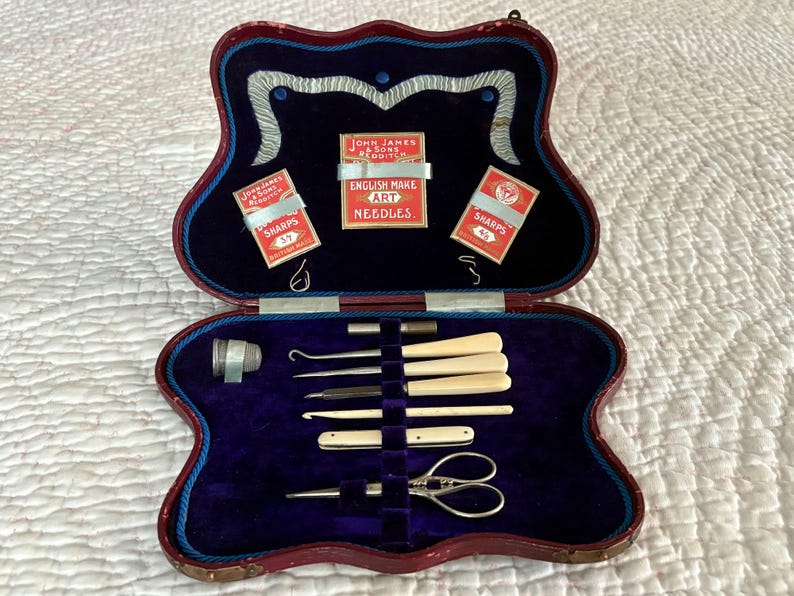 May include: An open antique sewing kit with a burgundy, scalloped case and a deep blue velvet interior. The kit contains sewing tools, including needles, hooks, and scissors. The case also holds three small boxes of needles with the text "SHARPS".