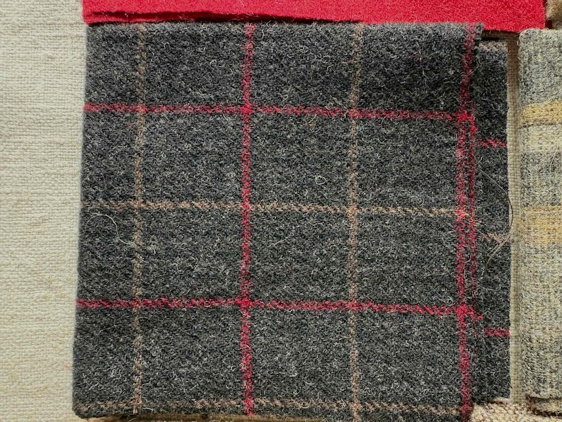 May include: Close-up of a gray tweed fabric with a red and tan plaid pattern. The fabric has a textured surface and is likely wool or a wool blend. A red fabric is at the top, and other fabric samples are visible.