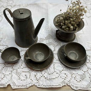 May include: Antique metal tea set with a teapot, creamer, two teacups, and saucers. A small decorative vase holds dried flowers. The set is displayed on a white lace tablecloth, creating a vintage aesthetic.