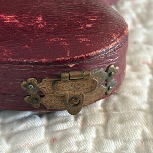 May include: Close-up of a burgundy-colored, antique jewelry box. The box has a distressed leather exterior and a decorative, ornate brass clasp. The box is resting on a white, textured surface.