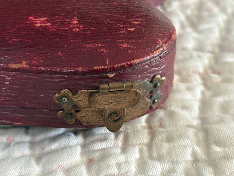 May include: Close-up of a burgundy-colored, antique jewelry box. The box has a distressed leather exterior and a decorative, ornate brass clasp. The box is resting on a white, textured surface.