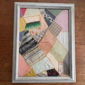 May include: A rectangular wall art piece featuring a patchwork of various fabric scraps in shades of pink, green, yellow, blue, and white, joined by gold-coloured stitching and enclosed in a distressed white wooden frame.