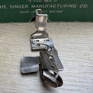 May include: A vintage Singer sewing machine attachment, made of silver-toned metal. The attachment has a curved shape with a flat base and a hinged, adjustable component. The text "THE SINGER MANUFACTURING CO. MADE IN GREAT BRITAIN" is visible.