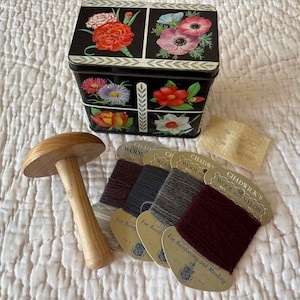 May include: A black floral sewing box with a wooden mushroom-shaped darning egg, spools of wool thread, and a small piece of fabric. The box is decorated with colorful flowers. The thread is in shades of gray and burgundy.
