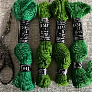 May include: Four spools of DMC embroidery floss in shades of green. The spools are labelled with the DMC numbers 911, 905, 905, and 70. Each spool has a label that reads "A Brode Qualite Special DMC 12" and includes the length in metres and yards. The spools are arranged in a row on a white linen fabric background.