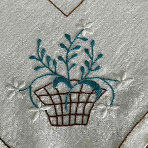 May include: White linen fabric with an embroidered design. The design features a brown basket filled with teal-coloured leaves and stems, and white flower accents. Brown stitching outlines the edges of the fabric.