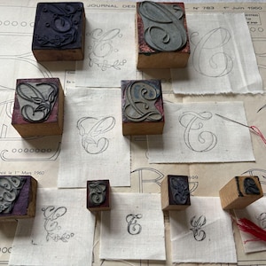 May include: A collection of wooden printing blocks featuring various designs, including letters and floral motifs. The blocks are arranged on a white surface with several pieces of fabric, each with a different design printed on it. The fabric pieces are labelled with the number 783 and the date 1st June 1960.
