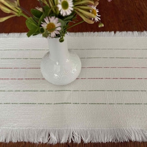 May include: A white woven table runner with a fringed edge, accented with thin red and green stripes. A white vase with a floral arrangement sits on the runner. The table runner is approximately 12 inches wide.