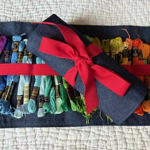 May include: A denim thread organiser with a red ribbon bow, holding spools of colourful embroidery floss. The floss colours range from pinks and blues to greens and oranges. The organiser is on a white quilted surface.