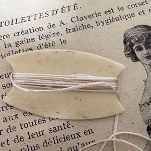 May include: A white bone thread bobbin with a beige thread wound around it. The bobbin is on a page of a book with French text about clothing.