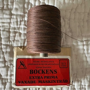 May include: A spool of brown waxed thread resting on a cardboard box with a red label. The label features the text Holma-Helsinglands Linspinneri & Vafveri Aktiebolag Forsa Sweden, Bockens, Extra Prima, Vaxade Maskintrad, and the number 25/3.