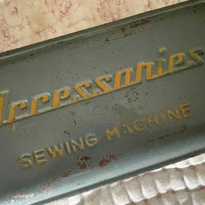 May include: A rectangular, weathered grey metal storage tin with embossed yellow lettering that reads Accessories and SEWING MACHINE. The surface shows signs of age, including minor rust spots and paint wear across the lid and edges.