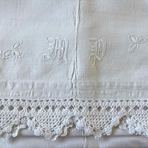 May include: Close-up of a white linen textile with embroidered floral details and initials. The bottom edge features a delicate, openwork lace trim. The fabric is divided by a vertical seam. The overall aesthetic is vintage and handcrafted.