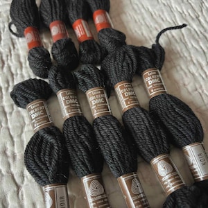 以下が含まれることがあります： 黒色の刺繍糸の束のクローズアップ。各束には「Laine Colbert DMC」と「NOIR」のラベルが付いています。糸はしっかりと巻かれており、ウールのように見えます。刺繍、クロスステッチ、その他の手芸プロジェクトに最適です。