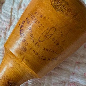 May include: A vintage wooden darning egg with a smooth, light brown finish. The egg-shaped tool has a handle and is stamped with text and a logo in dark red ink. The text includes the words "DARNING" and "WOOLLEN ARTICLES".