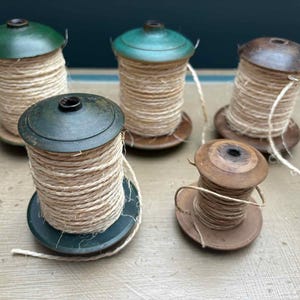 May include: Five vintage twine spools with wooden bases and lids in green and brown hues. The twine is a natural beige colour, with a length extending from each spool. The spools are arranged on a light-coloured surface. The spools are a charming, rustic item.