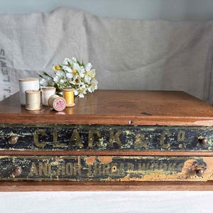 May include: A wooden two-drawer storage cabinet with distressed black and gold labels reading CLARK & CO and ANCHOR THREAD WORKS. Several small wooden spools of thread in white, beige, and yellow sit on top next to a small cluster of white flowers.