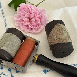 May include: A vintage printing roller set with a black handle, a metal frame, and two rollers. One roller is orange, and the other is dark brown with a floral design. A pink rose and a white and blue patterned cloth are in the background.
