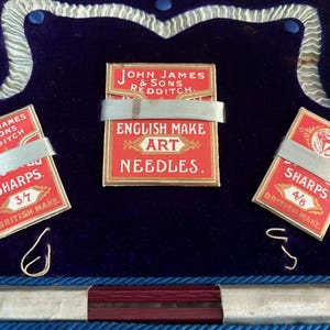 May include: An open box with a deep blue velvet interior displays three small red and gold needle packets. The packets are labeled "SHARPS" and "ENGLISH MAKE ART NEEDLES." The text "JOHN JAMES & SONS REDDITCH" is also visible.