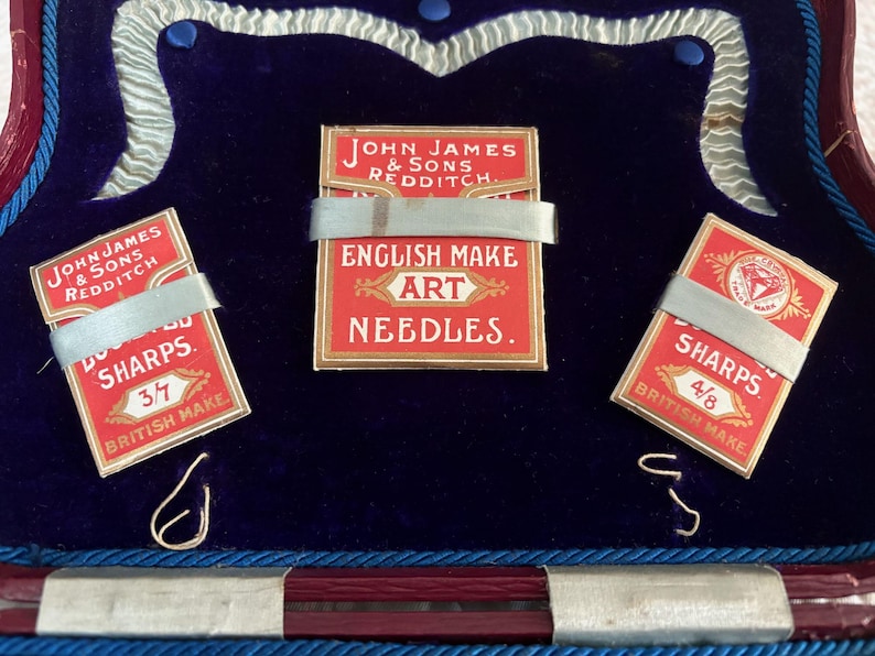 May include: An open box with a deep blue velvet interior displays three small red and gold needle packets. The packets are labeled "SHARPS" and "ENGLISH MAKE ART NEEDLES." The text "JOHN JAMES & SONS REDDITCH" is also visible.