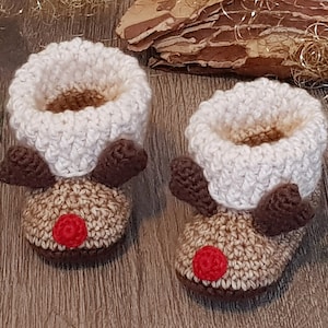 Könnte beinhalten: Ein Paar gehäkelte Babyschuhe in Form von Rentieren. Die Schuhe sind braun und beige mit weißen Bündchen und roten Bommeln als Nasen.